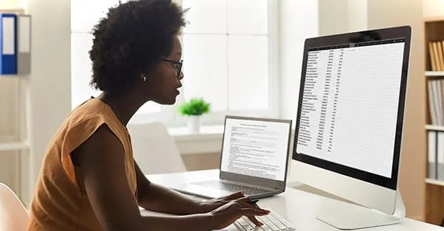 Mujer mirando la pantalla de una computadora de escritorio comparando dos listas.