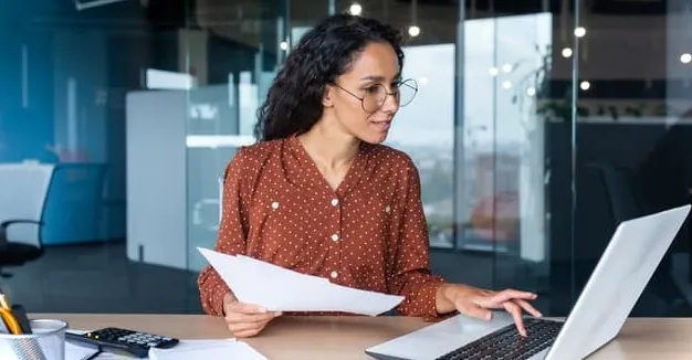 Mujer sosteniendo papeleo y mirando una computadora portátil mientras aprende a crear un diagrama de puntos simple en Excel