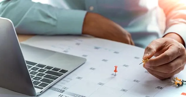 Hombre tomando notas en un calendario grande debajo de una computadora portátil abierta